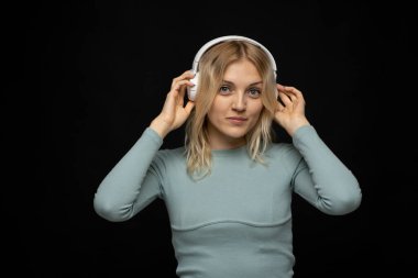 Beyaz kulaklıkla müzik dinleyen ve siyah arka plandaki kameraya bakan mutlu sarışın kız. Çekici kız eğleniyor ve hayattan zevk alıyor. Çekici kadın, en sevdiği şarkıyı seviyor.