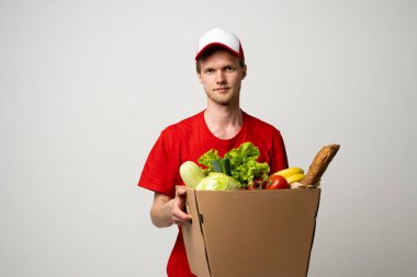 Kırmızı üniformalı teslimatçı yiyecek, meyve ve sebzelerle uğraşıyor. Postacı ve market teslimat servisi.