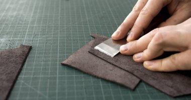 Bir deri işçisinin elleri, kahverengi deri parçalarını kesme hasırında inceltmek için metal bir kayak aleti kullanıyor. Görüntü, kaliteli deri ürünler üretmek için gereken hassasiyeti ve inceliği yakalıyor..