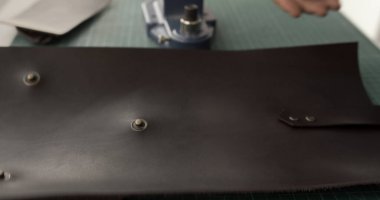 Deri bir parçanın kısmen odaklanmış bir görüntüsü kesme paspasına yerleştirilmiş kopçaları var. El yapımı bir nesnenin sürmekte olan montaj sürecini işaret eden bir el sıkışma makinesi arka planda görünür.