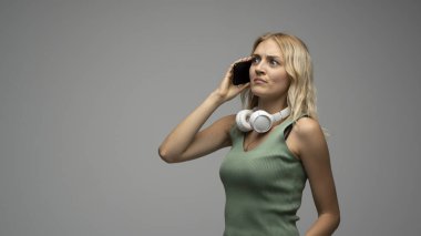 Boynuna kablosuz kulaklık takmış, kulağına akıllı telefon dayamış, kafası karışmış ya da endişeli gri arka planda izole edilmiş genç sarışın bir kadının portresi..