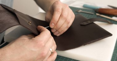 Esnaf ellerinin, kahverengi deri bir parçaya iğneyi ve ipliği aletlerle dikkatle bağladığı detaylı görüntü.