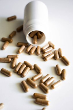 Kahverengi kapsüller etrafa saçılmış beyaz bir takviye şişesi. Doğal katkı maddeleri ve dozaj kontrolünü vurgulayan, sağlıkla ilgili bir natürmort konsepti.