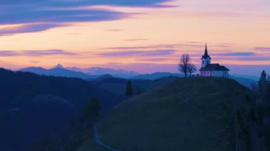Tepenin tepesinde St. Jacob Katolik kilisesi ve günbatımından sonra Alpler olan Slovenya 'nın güzel ve sakin manzarası. Turizm, tatil, yürüyüş ve doğa kavramları - 4K