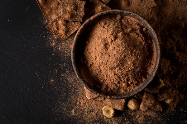 stock image Broken chocolate nuts pieces and cocoa powder in bowl on dark stone background