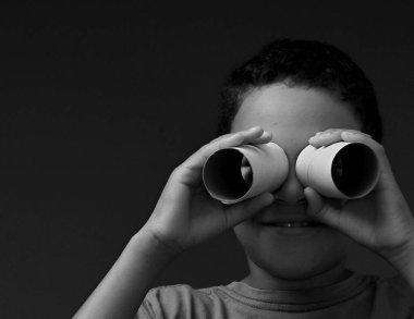 Gri arka planda tuvalet kağıdı rulolarından yapılmış dürbünlere bakan çocuk.