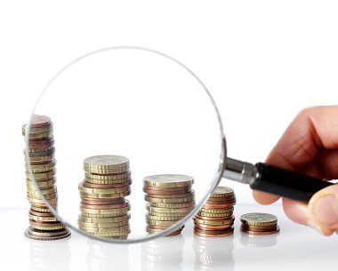 Beyaz arka plandaki madeni para yığınlarının üzerinde büyüteç tutan el.