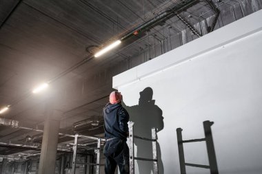 Miğfer takan erkek bir inşaat işçisi büyük bir sanayi deposunda duvar boyuyor.