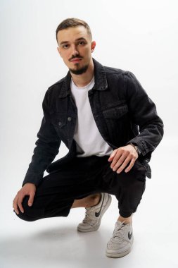 Siyah ceketli, siyah pantolonlu ve gri stüdyo arka planında beyaz tişörtlü yakışıklı bir adam.