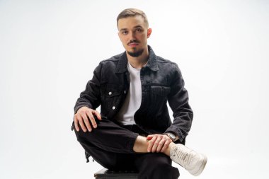 Siyah ceketli, siyah pantolonlu ve gri stüdyo arka planında beyaz tişörtlü yakışıklı bir adam.