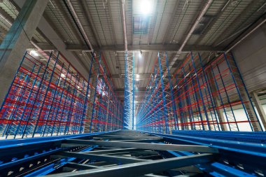 Siyah elbiseli bir erkek mimar, uzun metal rafları olan boş bir ticari depoda telefonla konuşuyor.