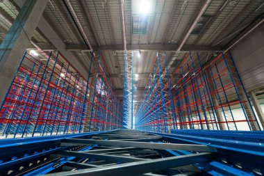 Siyah elbiseli bir erkek mimar, uzun metal rafları olan boş bir ticari depoda telefonla konuşuyor.