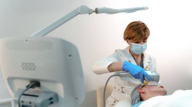 Bir güzellik uzmanı, modern bir güzellik kliniğinde, yetişkin bir kadının yüzünde lazer makinesi kullanır.