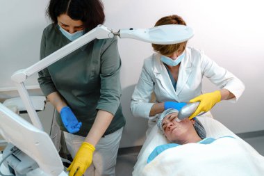 Koruyucu maskeli ve eldivenli iki sağlık uzmanı, klinik bir ortamda gelişmiş cilt teknolojisi kullanarak, bir hastaya zararsız yüz derisi tedavisi uyguluyor.