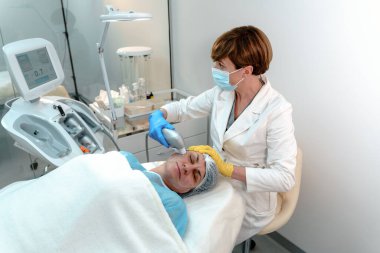 Bir güzellik uzmanı, modern bir güzellik kliniğinde, yetişkin bir kadının yüzünde lazer makinesi kullanır.