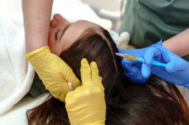 Genç bir kadının kafa derisinde saç büyümesi için mezoterapi uygulayan iki profesyonelin yakın çekimi klinik bir ortamda hassasiyet ve bakım sağlar.