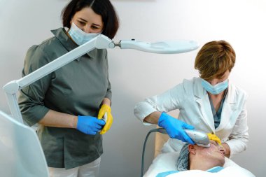 Koruyucu maskeli ve eldivenli iki sağlık uzmanı, klinik bir ortamda gelişmiş cilt teknolojisi kullanarak, bir hastaya zararsız yüz derisi tedavisi uyguluyor.