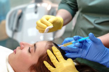 Genç bir kadının kafa derisinde saç büyümesi için mezoterapi uygulayan iki profesyonelin yakın çekimi klinik bir ortamda hassasiyet ve bakım sağlar.