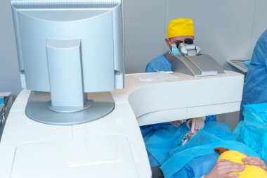 A surgeon in a yellow cap operates a microscope, assisted by a monitor, during a precision eye surgery. Sterile blue drapes cover the patient, ensuring a controlled environment clipart