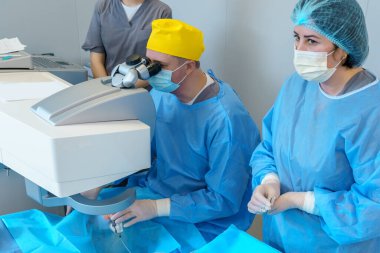 Koruyucu kıyafetli cerrahi ekip özel bir mikroskop kullanan bir cerrah tarafından yapılan lazer göz ameliyatını yönetiyor. Ekipman ve steril kurulum hastanın güvenliğini sağlar