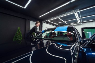 Kadın teknisyen titizlikle arabanın ön camını mikrofiber bir bez kullanarak temizliyor. Seri olmayan, lekesiz bir son sağlıyor.