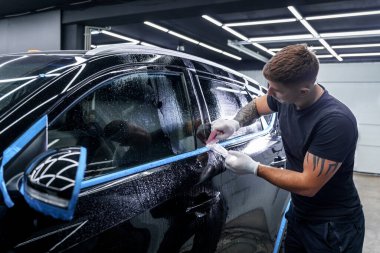 Dövmeli bir adam araba tamirhanesindeki siyah bir arabaya koruyucu film uyguluyor.