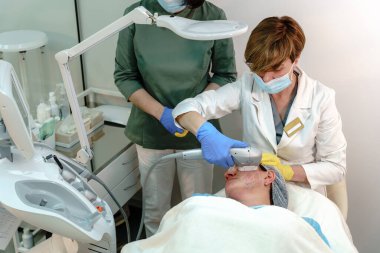 Koruyucu maskeli ve eldivenli iki sağlık uzmanı, klinik bir ortamda gelişmiş cilt teknolojisi kullanarak, bir hastaya zararsız yüz derisi tedavisi uyguluyor.