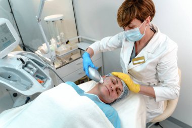 Bir güzellik uzmanı, modern bir güzellik kliniğinde, yetişkin bir kadının yüzünde lazer makinesi kullanır.