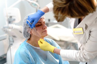 Estetik uzmanı, klinik bir ortamda olgun bir kadına yüz haritası yapar. Hedeflenmiş cilt bakımı için hassas bir şekilde hazırlanır.