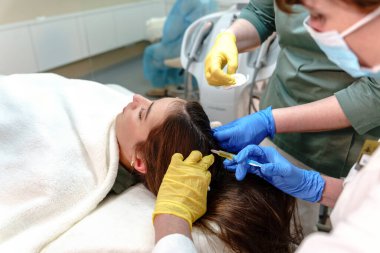 Genç bir kadının kafa derisinde saç büyümesi için mezoterapi uygulayan iki profesyonelin yakın çekimi klinik bir ortamda hassasiyet ve bakım sağlar.