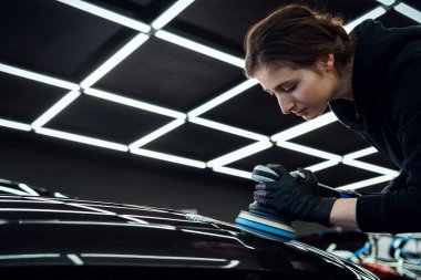 Kendini işine adamış bir kadın işçi, profesyonel bir otomotiv atölyesindeki kusurları gidermek ve parlaklığı artırmak için bir araba yüzeyini dikkatlice parlatır.