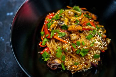 Karışık deniz ürünleri, renkli sebzeler ve susam tohumlu, Asya 'dan esinlenilmiş canlı bir yemek için taze otlarla süslenmiş lezzetli bir kase kızarmış cam erişte.