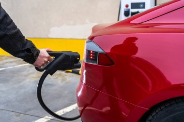 Birisi kırmızı elektrikli bir arabaya şarj kablosu takıyor, çevre dostu teknolojiyi ve modern ulaşım için sürdürülebilir enerji çözümlerini vurguluyor