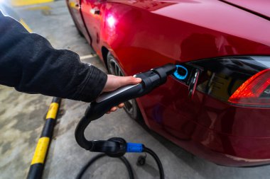 Birisi kırmızı elektrikli bir arabaya şarj kablosu takıyor, çevre dostu teknolojiyi ve modern ulaşım için sürdürülebilir enerji çözümlerini vurguluyor
