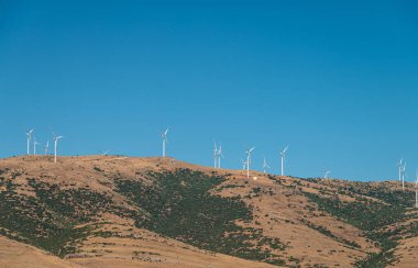 Rüzgar türbinleri tepede duruyor ve güneşli mavi gökyüzünde elektrik üretiyorlar.