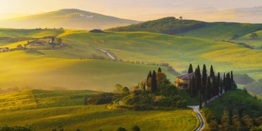 İtalya, San Quirico d 'Orcia' da gündoğumunda Val d 'Orcia vadisinin sisli sabah güneşli tepeleri arasında selvi ağaçlarıyla çevrili bir ev.