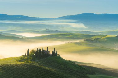 Pienza, Tuscany, İtalya - 5 Mayıs 2023: Ev, San Quirico d 'Orcia, İtalya' daki Val d 'Orcia vadisinin puslu sabah güneşli tepeleri arasında selvi ağaçlarıyla çevrili.