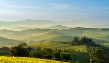 İtalya, San Quirico d 'Orcia' da gündoğumunda Val d 'Orcia vadisinin sisli sabah güneşli tepeleri arasında selvi ağaçlarıyla çevrili bir ev.