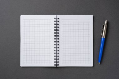 Yanı başında tükenmez kalem olan koyu gri bir masanın üzerinde işaretli ve sarmal bir defter.