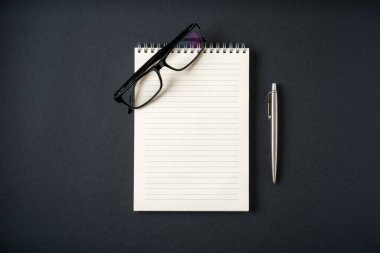 Koyu gri arkaplanda defter, gözlük ve tükenmez kalem üst görünümü