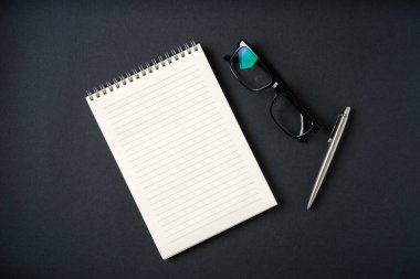Koyu gri arkaplanda defter, gözlük ve tükenmez kalem üst görünümü