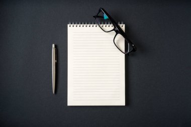 Koyu gri arkaplanda defter, gözlük ve tükenmez kalem üst görünümü