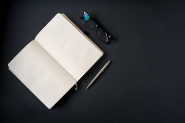 Koyu gri arkaplanda defter, gözlük ve tükenmez kalem üst görünümü