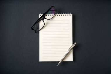 Koyu gri arkaplanda defter, gözlük ve tükenmez kalem üst görünümü