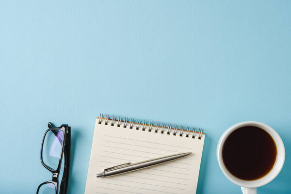 Top view of notebook, glasses, coffee and ballpoint pen on blue background