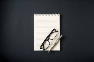 Koyu gri arkaplanda defter, gözlük ve tükenmez kalem üst görünümü