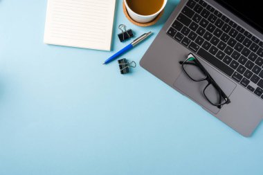 Kahve fincanı, dizüstü bilgisayar klavyesi ve mavi ofis masasında çeşitli kırtasiye malzemeleri. İş ofisi kavramı