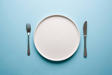 Mavi arkaplanda boş seramik beyaz tabak ve çatal bıçak takımlarının üst görünümü