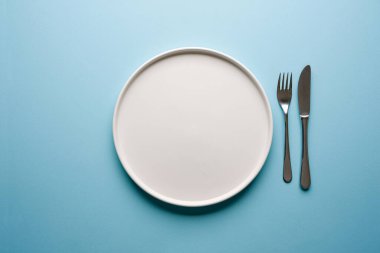 Mavi arkaplanda boş seramik beyaz tabak ve çatal bıçak takımlarının üst görünümü