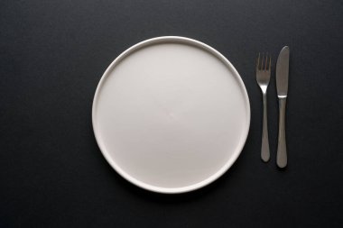 Boş seramik beyaz tabak ve siyah zemin üzerindeki çatal bıçak takımlarının üst görünümü